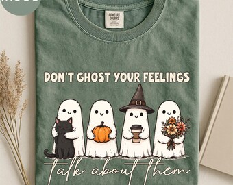 May include: A moss green t-shirt featuring the text "DON'T GHOST YOUR FEELINGS" and "talk about them" with a graphic of four white ghosts. One ghost holds a black cat, one holds an orange pumpkin, one wears a witch hat holding a coffee cup, and one holds a bouquet of flowers.