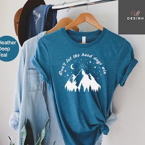 Pu&ograve; includere: T-shirt color verde acqua con la scritta "Don't let the hard days win" sopra una grafica di montagna e foresta. La maglietta &egrave; appesa a una gruccia di legno, con altri capi sullo sfondo.