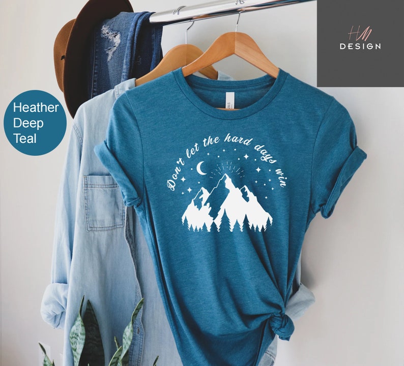 Pu&ograve; includere: T-shirt color verde acqua con la scritta "Don't let the hard days win" sopra una grafica di montagna e foresta. La maglietta &egrave; appesa a una gruccia di legno, con altri capi sullo sfondo.