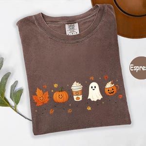 K&ouml;nnte beinhalten: Ein espressofarbenes T-Shirt mit Rundhalsausschnitt und einem Comfort Colors Markenetikett. Das Shirt zeigt einen Grafikdruck mit einem l&auml;chelnden orangefarbenen Ahornblatt, einem K&uuml;rbis, einem Kaffeebecher, einem wei&szlig;en Geist und einer K&uuml;rbistasse.