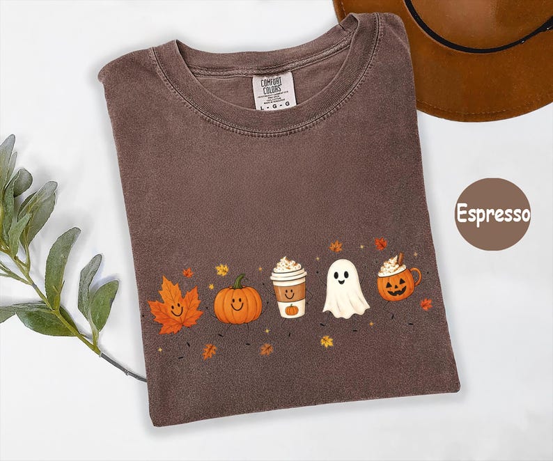 K&ouml;nnte beinhalten: Ein espressofarbenes T-Shirt mit Rundhalsausschnitt und einem Comfort Colors Markenetikett. Das Shirt zeigt einen Grafikdruck mit einem l&auml;chelnden orangefarbenen Ahornblatt, einem K&uuml;rbis, einem Kaffeebecher, einem wei&szlig;en Geist und einer K&uuml;rbistasse.