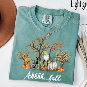 May include: Light green t-shirt with a fall-themed graphic. The design features a gnome, pumpkins, a black cat, and autumn trees. The text "Ahhhh...fall" is written in a cursive font. A silver necklace is draped across the shirt.