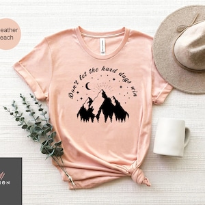 Pu&ograve; includere: T-shirt color pesca con grafica di montagna e la scritta "Don't let the hard days win". Il design include una luna, stelle e un sole. La maglietta &egrave; abbinata a un cappello neutro e una tazza bianca.