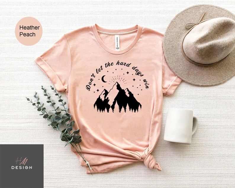 Pu&ograve; includere: T-shirt color pesca con grafica di montagna e la scritta "Don't let the hard days win". Il design include una luna, stelle e un sole. La maglietta &egrave; abbinata a un cappello neutro e una tazza bianca.