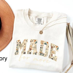 May include: Ivory-colored t-shirt with the word "MADE" in a floral pattern, and the words "for more" in a script font. A gold necklace with a pendant is on the shirt. The shirt is folded on a white surface.