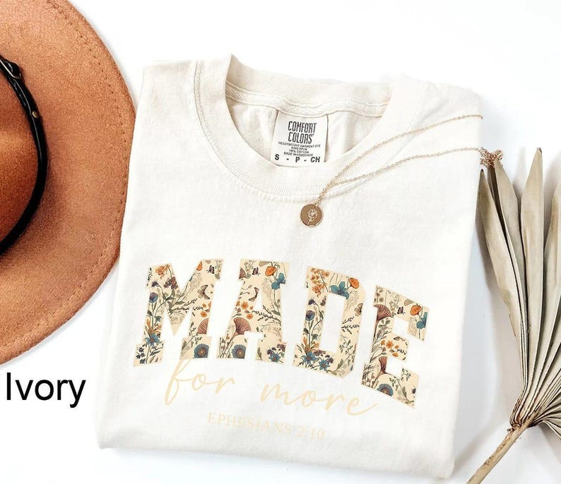 May include: Ivory-colored t-shirt with the word "MADE" in a floral pattern, and the words "for more" in a script font. A gold necklace with a pendant is on the shirt. The shirt is folded on a white surface.