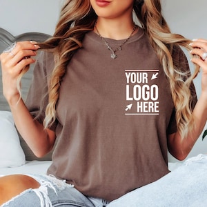 May include: A brown short-sleeved t-shirt featuring white text on the left chest area that reads "YOUR LOGO HERE" with two white arrows pointing toward the text. The shirt has a crew neckline and is worn with light blue distressed denim jeans.