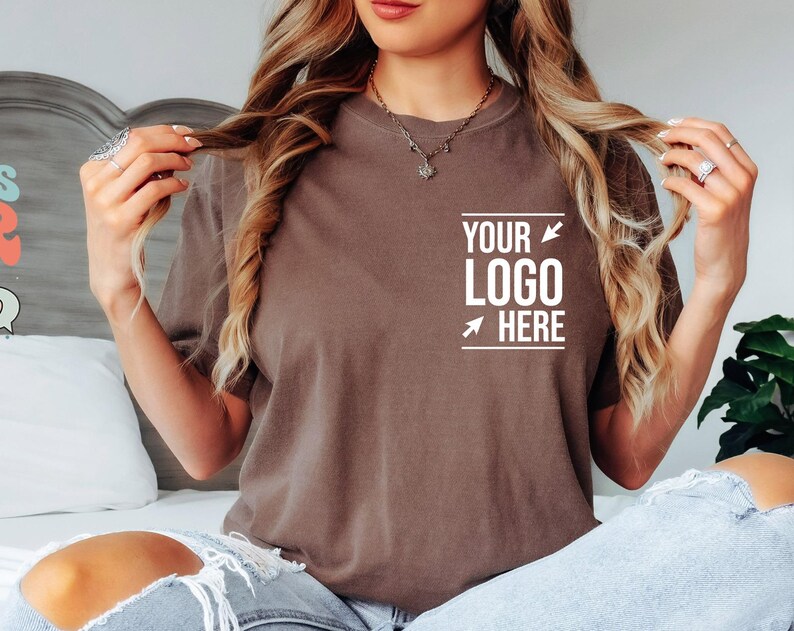 May include: A brown short-sleeved t-shirt featuring white text on the left chest area that reads "YOUR LOGO HERE" with two white arrows pointing toward the text. The shirt has a crew neckline and is worn with light blue distressed denim jeans.