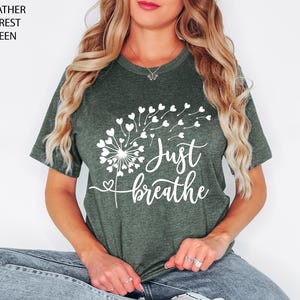 以下が含まれることがあります： ヘザーフォレストグリーンの半袖Tシャツ。ハート型の種が風に舞うタンポポの白いグラフィックが描かれています。タンポポのデザインの下には、白い筆記体で「Just breathe」という文字がプリントされています。