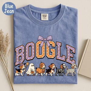 K&ouml;nnte beinhalten: Ein blaues T-Shirt mit dem Schriftzug BOOGLE in orangefarbenen und violetten gepunkteten Buchstaben und einer violetten Schleife dar&uuml;ber. Darunter befinden sich sechs Beagle-Hunde in Halloween-Kost&uuml;men: Hexe, Geist, K&uuml;rbis, Vampir, Skelett und Mumie.