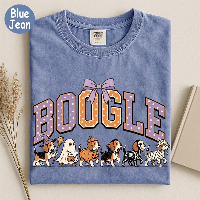 K&ouml;nnte beinhalten: Ein blaues T-Shirt mit dem Schriftzug BOOGLE in orangefarbenen und violetten gepunkteten Buchstaben und einer violetten Schleife dar&uuml;ber. Darunter befinden sich sechs Beagle-Hunde in Halloween-Kost&uuml;men: Hexe, Geist, K&uuml;rbis, Vampir, Skelett und Mumie.