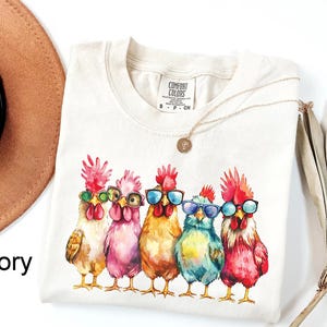 Puede incluir: Una camiseta de color crema con una ilustraci&oacute;n de acuarela de cinco pollos con gafas de sol. Los pollos son de color rosa, azul, amarillo y blanco. El texto "COMFORT COLORS" est&aacute; impreso en la camiseta.