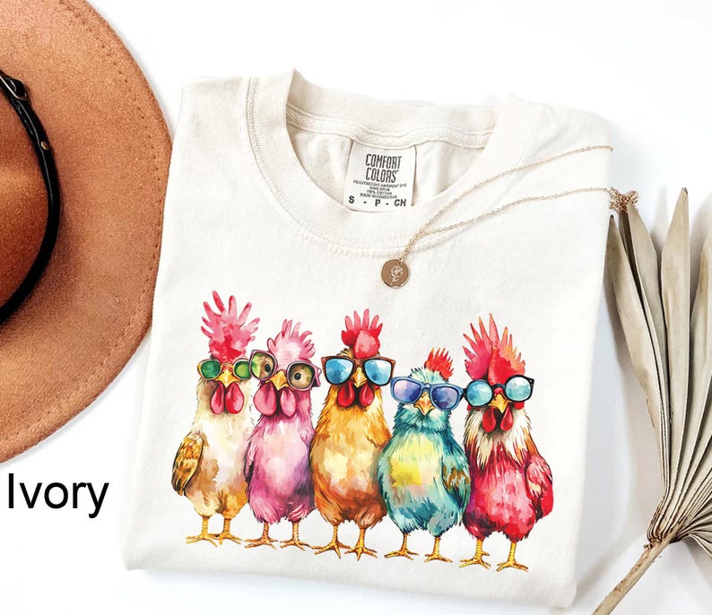 Puede incluir: Una camiseta de color crema con una ilustraci&oacute;n de acuarela de cinco pollos con gafas de sol. Los pollos son de color rosa, azul, amarillo y blanco. El texto "COMFORT COLORS" est&aacute; impreso en la camiseta.