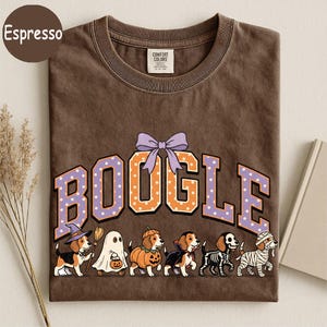 K&ouml;nnte beinhalten: Ein espressobraunes T-Shirt mit dem Schriftzug BOOGLE in gro&szlig;en orangefarbenen und violetten gepunkteten Buchstaben, gekr&ouml;nt von einer violetten Schleife. Darunter befinden sich sechs Beagle-Hunde in Halloween-Kost&uuml;men: Hexe, Geist, K&uuml;rbis, Vampir, Skelett und Mumie.