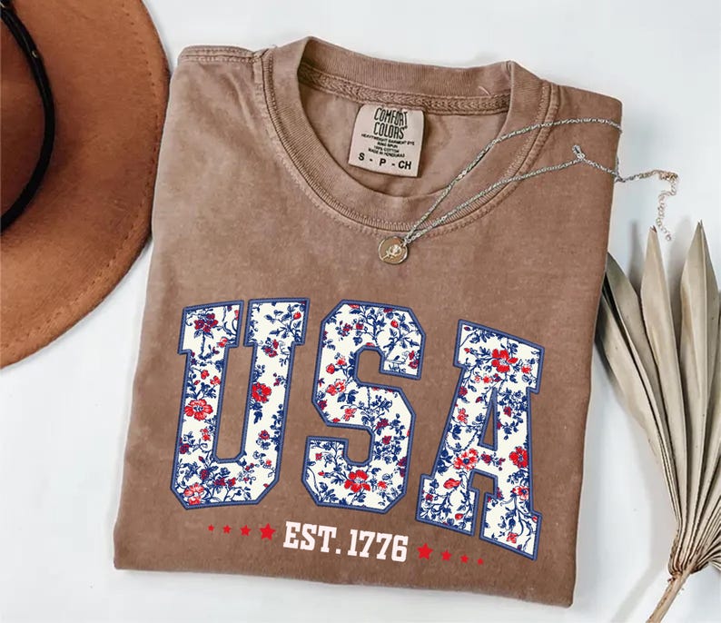 May include: A brown Comfort Colors t-shirt with the word "USA" in a floral pattern. The shirt also features the text "EST. 1776" in white. A silver necklace with a pendant is draped across the shirt.