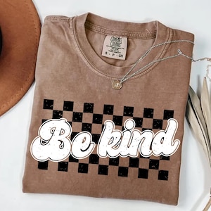 Pu&ograve; includere: Una t-shirt a maniche corte color marrone chiaro con scollo rotondo, caratterizzata da una grafica a scacchi neri e marroni sul petto. Il testo "Be kind" &egrave; stampato in un carattere bianco, arrotondato e dall'aspetto vissuto sopra il motivo a scacchi. La maglietta presenta l'etichetta del marchio Comfort Colors all'interno del colletto.