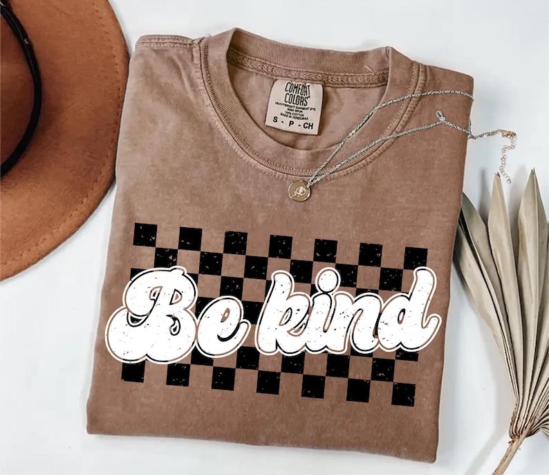 Pu&ograve; includere: Una t-shirt a maniche corte color marrone chiaro con scollo rotondo, caratterizzata da una grafica a scacchi neri e marroni sul petto. Il testo "Be kind" &egrave; stampato in un carattere bianco, arrotondato e dall'aspetto vissuto sopra il motivo a scacchi. La maglietta presenta l'etichetta del marchio Comfort Colors all'interno del colletto.