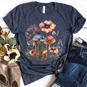 May include: A navy blue t-shirt featuring a watercolor illustration of colorful mushrooms and flowers. The design includes mushrooms with purple, blue, and red caps, surrounded by foliage and a large orange flower. The shirt is paired with blue jeans.
