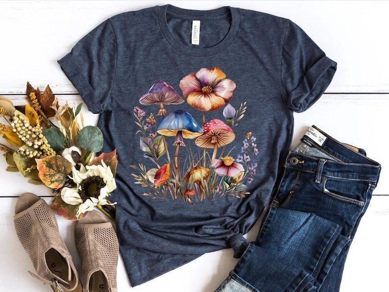 May include: A navy blue t-shirt featuring a watercolor illustration of colorful mushrooms and flowers. The design includes mushrooms with purple, blue, and red caps, surrounded by foliage and a large orange flower. The shirt is paired with blue jeans.
