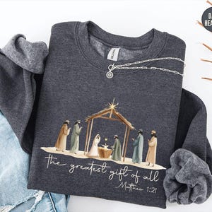 May include: Dark heather gray sweatshirt featuring a Nativity scene illustration. The graphic includes figures in robes, a wooden structure, and the text "the greatest gift of all" and "Matthew 1:21". A silver necklace with a pendant is draped on the sweatshirt.