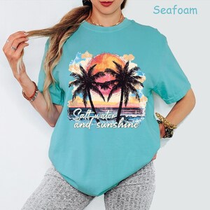 May include: Seafoam green t-shirt featuring a sunset scene with palm trees and the text "Salt water and sunshine." The graphic includes a vibrant orange sun, blue water, and colorful clouds. The shirt is a relaxed fit.