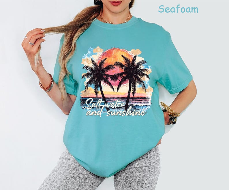May include: Seafoam green t-shirt featuring a sunset scene with palm trees and the text "Salt water and sunshine." The graphic includes a vibrant orange sun, blue water, and colorful clouds. The shirt is a relaxed fit.