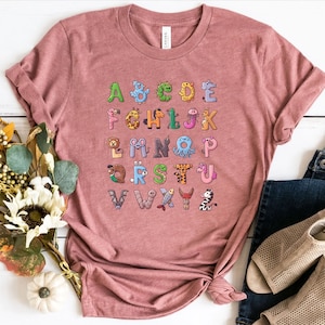 May include: A dusty rose-colored t-shirt featuring a colorful alphabet design. Each letter is creatively illustrated with an animal or creature, creating a fun and educational graphic. The shirt is styled with rolled-up sleeves and a casual knot.