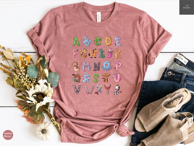 May include: A dusty rose-colored t-shirt featuring a colorful alphabet design. Each letter is creatively illustrated with an animal or creature, creating a fun and educational graphic. The shirt is styled with rolled-up sleeves and a casual knot.