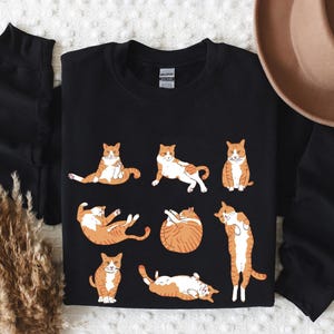 May include: A black long-sleeve sweatshirt featuring a graphic print of seven orange and white tabby cats in various poses, including sitting, lying down, and stretching. The garment has a crew neckline and a standard fit with ribbed cuffs.