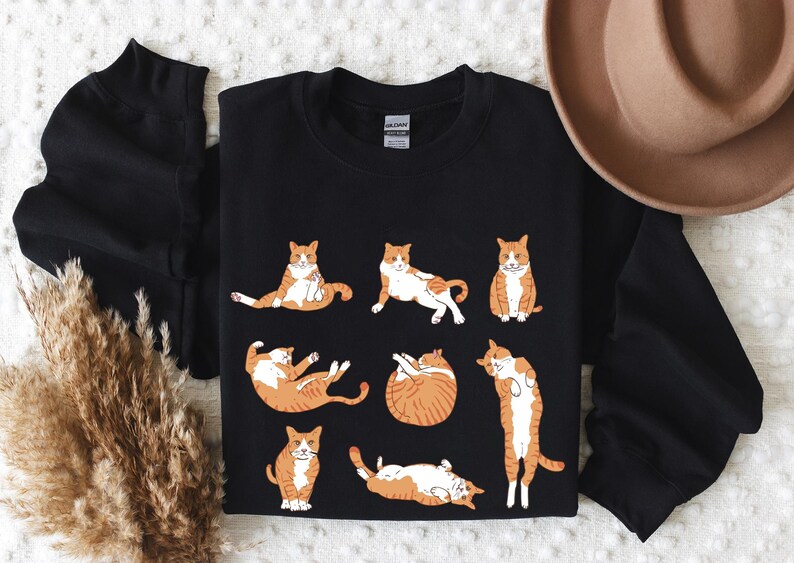 May include: A black long-sleeve sweatshirt featuring a graphic print of seven orange and white tabby cats in various poses, including sitting, lying down, and stretching. The garment has a crew neckline and a standard fit with ribbed cuffs.