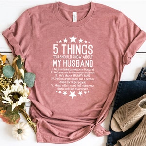 May include: A dusty rose t-shirt with the text "5 THINGS YOU SHOULD KNOW ABOUT MY HUSBAND". The text is in white and includes a list of characteristics. The shirt is folded and tied at the bottom, with a pair of jeans and shoes in the background.