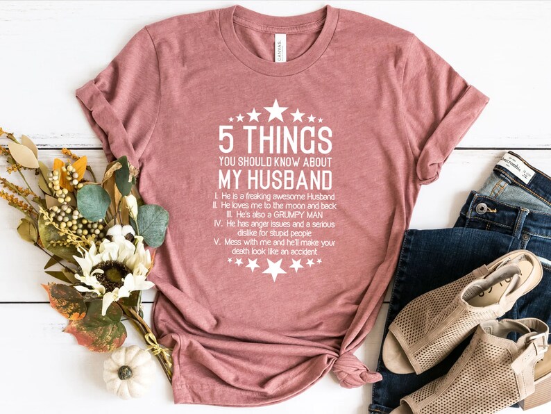 May include: A dusty rose t-shirt with the text "5 THINGS YOU SHOULD KNOW ABOUT MY HUSBAND". The text is in white and includes a list of characteristics. The shirt is folded and tied at the bottom, with a pair of jeans and shoes in the background.