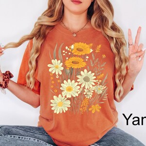 K&ouml;nnte beinhalten: Orangefarbenes T-Shirt mit einem Blumendesign mit gelben und wei&szlig;en G&auml;nsebl&uuml;mchen und Sonnenblumen. Die Grafik enth&auml;lt gr&uuml;ne Bl&auml;tter und Stiele. Das Wort "Yam" ist in Schwarz unten rechts auf dem T-Shirt aufgedruckt.