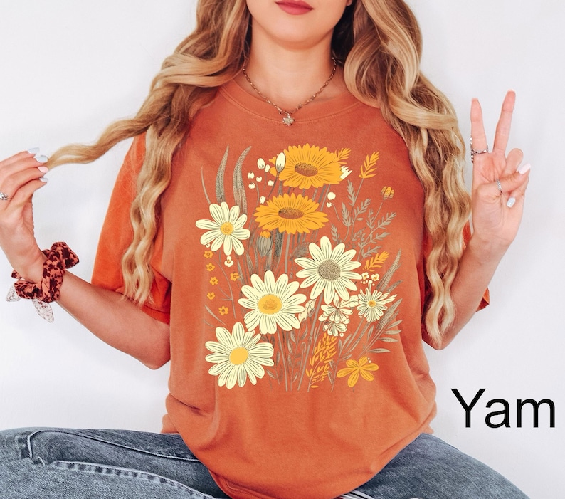 K&ouml;nnte beinhalten: Orangefarbenes T-Shirt mit einem Blumendesign mit gelben und wei&szlig;en G&auml;nsebl&uuml;mchen und Sonnenblumen. Die Grafik enth&auml;lt gr&uuml;ne Bl&auml;tter und Stiele. Das Wort "Yam" ist in Schwarz unten rechts auf dem T-Shirt aufgedruckt.
