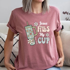 K&ouml;nnte beinhalten: Mauvefarbenes T-Shirt mit dem Text "Jesus Fills My Cup" und einer floralen Bechergrafik. Der Becher ist hellgr&uuml;n mit G&auml;nsebl&uuml;mchen-Akzenten. Das Shirt zeigt auch den Bibelvers Psalm 23:5.