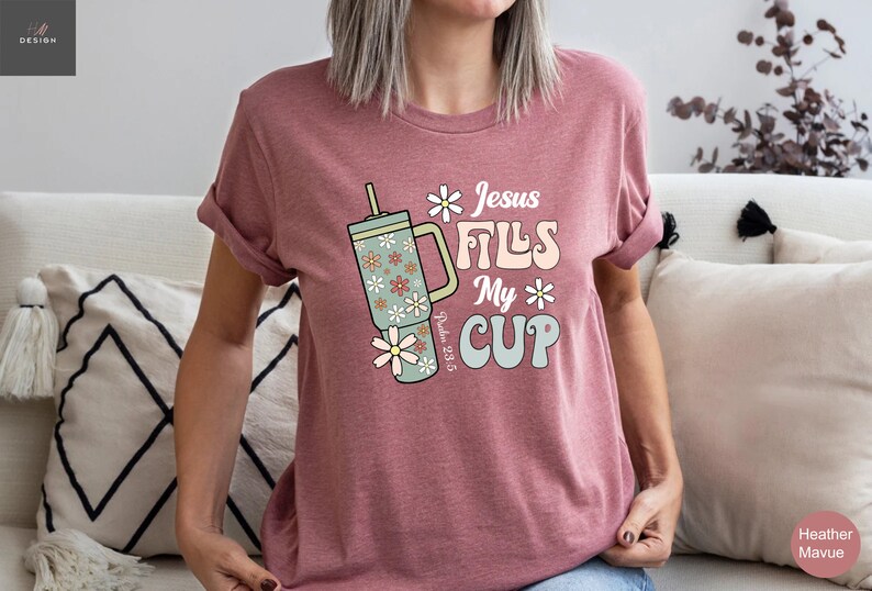 K&ouml;nnte beinhalten: Mauvefarbenes T-Shirt mit dem Text "Jesus Fills My Cup" und einer floralen Bechergrafik. Der Becher ist hellgr&uuml;n mit G&auml;nsebl&uuml;mchen-Akzenten. Das Shirt zeigt auch den Bibelvers Psalm 23:5.