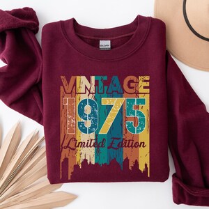 May include: A maroon sweatshirt with rolled-up sleeves. The front features the text "VINTAGE 1975 Limited Edition" in a retro color scheme of orange, teal, and yellow. A tan hat is visible in the upper right corner, and a decorative fan is in the lower left.