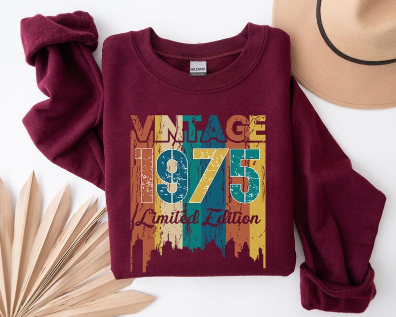 May include: A maroon sweatshirt with rolled-up sleeves. The front features the text "VINTAGE 1975 Limited Edition" in a retro color scheme of orange, teal, and yellow. A tan hat is visible in the upper right corner, and a decorative fan is in the lower left.