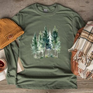May include: Heather military green long-sleeve shirt featuring a watercolor illustration of evergreen trees. The shirt is paired with a mustard-colored beanie, a plaid scarf, and a leather bag. The shirt's color is labeled as "Heather Military Green".