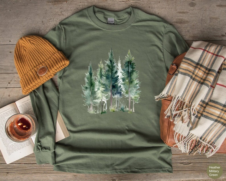 May include: Heather military green long-sleeve shirt featuring a watercolor illustration of evergreen trees. The shirt is paired with a mustard-colored beanie, a plaid scarf, and a leather bag. The shirt's color is labeled as "Heather Military Green".