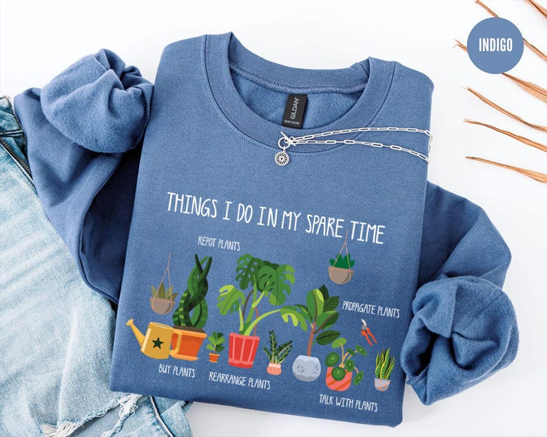 May include: Indigo blue sweatshirt with the text "THINGS I DO IN MY SPARE TIME" and illustrations of plants. The text includes phrases like "Repot Plants" and "Talk with Plants". A silver necklace is draped across the sweatshirt.