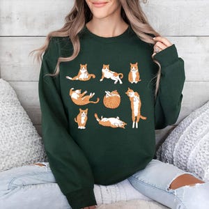 May include: A forest green long-sleeve crewneck sweatshirt featuring a graphic print of eight orange and white tabby cats in various poses, including sitting, stretching, and lying down, arranged in a grid pattern across the chest.