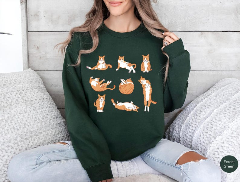 May include: A forest green long-sleeve crewneck sweatshirt featuring a graphic print of eight orange and white tabby cats in various poses, including sitting, stretching, and lying down, arranged in a grid pattern across the chest.