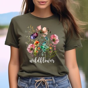 May include: Olive green t-shirt featuring a watercolor floral design with pink, purple, and blue flowers. The word "wildflower" is printed in white script below the flowers. The shirt is a crew neck style.
