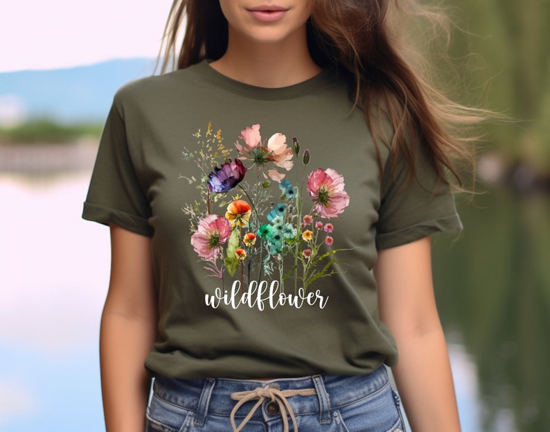 May include: Olive green t-shirt featuring a watercolor floral design with pink, purple, and blue flowers. The word "wildflower" is printed in white script below the flowers. The shirt is a crew neck style.