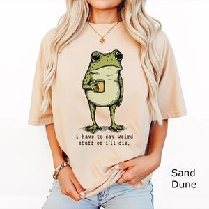 May include: A sand-coloured t-shirt featuring a green frog illustration holding a small mug. Below the frog, the text reads: i have to say weird stuff or i'll die. The shirt has short sleeves and a crew neckline, presented in a solid light tan colour.