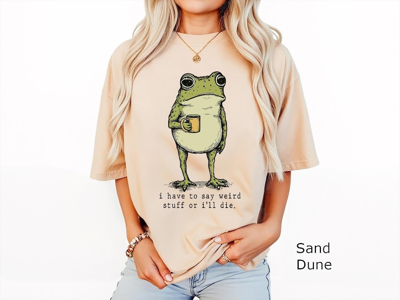 May include: A sand-coloured t-shirt featuring a green frog illustration holding a small mug. Below the frog, the text reads: i have to say weird stuff or i'll die. The shirt has short sleeves and a crew neckline, presented in a solid light tan colour.
