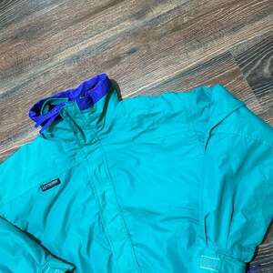 Könnte beinhalten: Eine tealblaue Jacke mit lila Futter und einem Columbia-Logo. Die Jacke hat einen Reißverschluss und eine Kapuze.