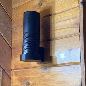 May include: A black cylindrical smart speaker mounted on a wooden wall. The speaker has a perforated lower section and a flat base. The wall is made of horizontal wooden planks with a natural wood finish.