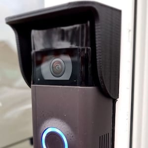 May include: A dark gray smart doorbell with a built-in camera and a black protective cover. The doorbell has a glowing blue ring around the button and the brand name "ring" is visible at the bottom.